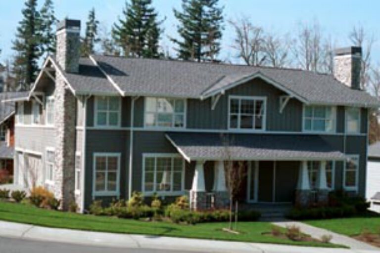 This is a two-story residential house with gray siding, white trim, stonework accents, multiple windows, and a well-manicured lawn on a sunny day.