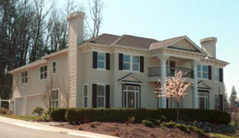 A two-story beige house with white trim, a prominent chimney, several windows, a garage door, a blossoming tree in front, and a clear blue sky.