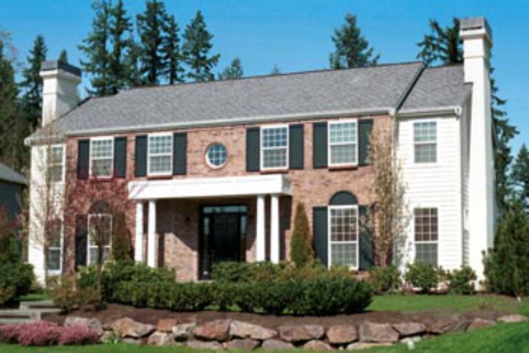 This is a two-story brick house with white trim, a central entrance, a manicured lawn, and surrounding trees under a clear blue sky.