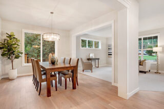 Bright, modern dining area with a wooden table, beige chairs, and a hanging pendant light. Large windows, greenery outside, adjacent sitting room visible.