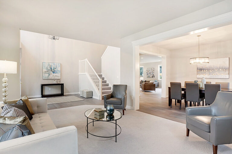 A spacious, well-lit living room with neutral colors features a fireplace, modern furniture, a staircase, and adjoining dining area with a hanging chandelier.