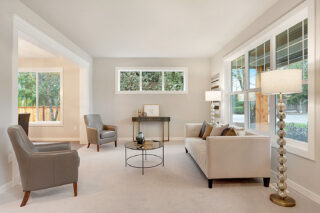 A bright living room with large windows, a white sofa, two armchairs, a glass coffee table, decorative lamps, and a view of trees outside.