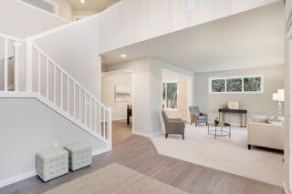 This image features a bright, spacious living room with a staircase, neutral colored furniture, hardwood floors, and a high ceiling.