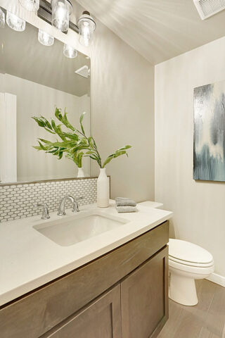 This image shows a modern bathroom with a white countertop sink, wooden cabinets, a toilet, artistic wall decor, and a plant accent.