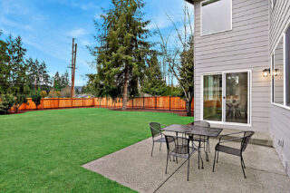 An image showing a well-kept backyard with a lawn, patio furniture, wooden fence, trees, and the exterior of a two-story modern house.