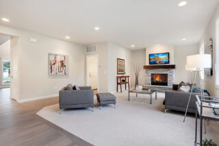 This image shows a spacious modern living room with a fireplace, grey furniture, hardwood floors, decorative artwork, and a mounted television.