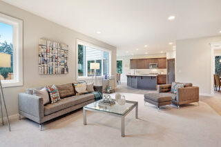 A modern living room with a gray sofa, armchair, ottoman, and glass coffee table. Open concept design leads to a kitchen with dark cabinets.