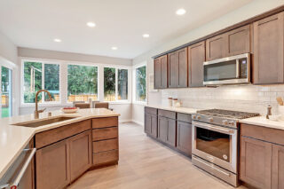 A modern kitchen with dark wood cabinets, stainless steel appliances, white countertops, subway tile backsplash, and a view of trees through large windows.