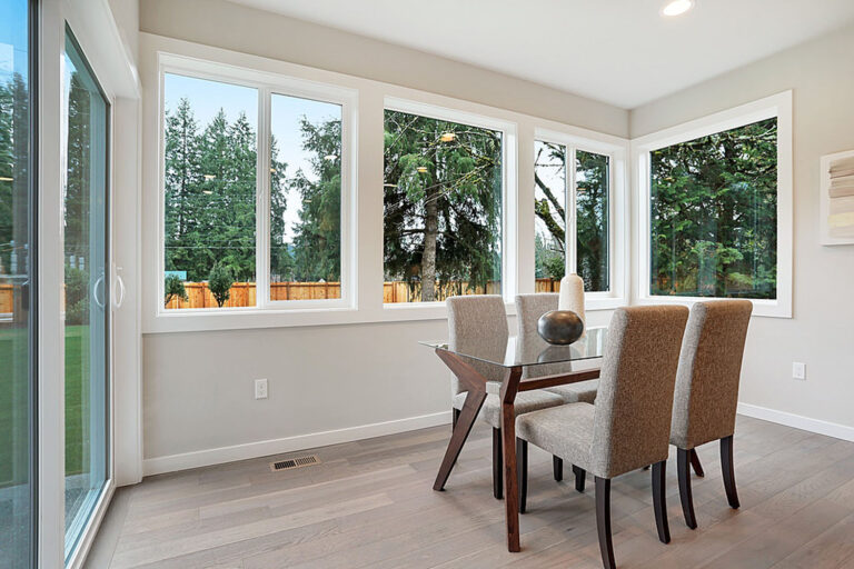 Bright dining room interior with large windows, a wooden table, four upholstered chairs, and a view of trees outside. Modern, clean, and well-lit space.