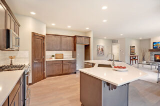 This is a modern kitchen with dark wood cabinets, stainless steel appliances, and a white countertop island. It opens into a living area with a fireplace.