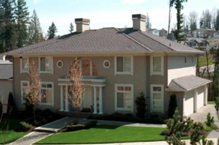 A two-story beige house with a gray roof features a well-manicured green lawn, a detached garage, and young trees in a suburban neighborhood.