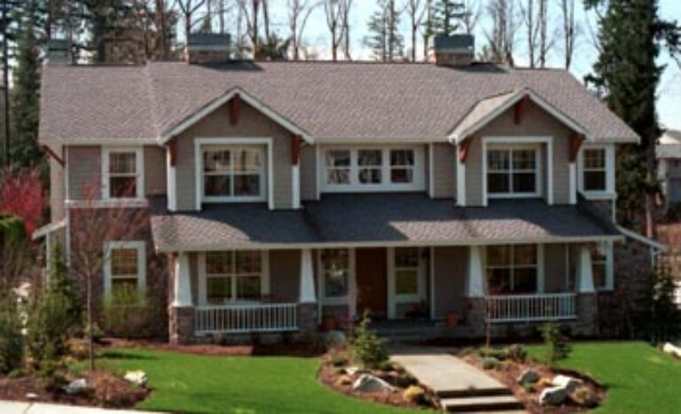  A two-story house with a reddish-brown roof, white trim, multiple windows, a front porch with pillars, surrounded by a manicured lawn and landscaping. 