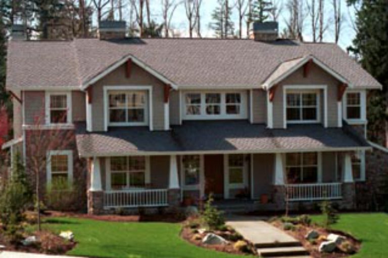 A two-story house with a reddish-brown roof, white trim, multiple windows, a front porch with pillars, surrounded by a manicured lawn and landscaping.