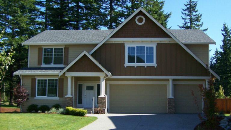 Two-story suburban house with brown siding, white trim, garage, and small lawn. Surrounded by trees under a clear sky. No people visible.