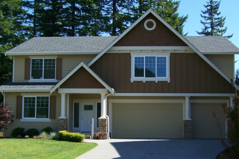 Two-story suburban house with brown siding, white trim, garage, and small lawn. Surrounded by trees under a clear sky. No people visible.