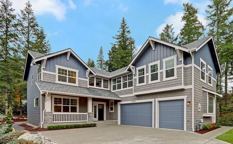  A two-story blue-gray house with a gabled roof, multiple windows, a double garage, and a small front porch, surrounded by trees under a cloudy sky. 