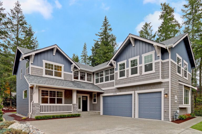 A two-story suburban house with gray siding and white trim, a two-car garage, and a porch, surrounded by trees under a cloudy sky.