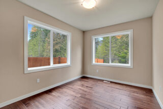 Empty room with tan walls, dark wood flooring, two large windows showing trees outside, and a single ceiling light fixture.