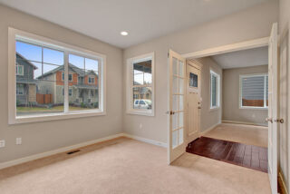 The image shows an empty room with beige carpeting, large windows, white walls, an open French door, and a view of neighboring houses outside.