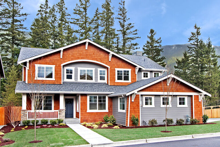 A two-story house with orange siding, gray accents, white windows, a green lawn, and pine trees in the background under a cloudy sky.