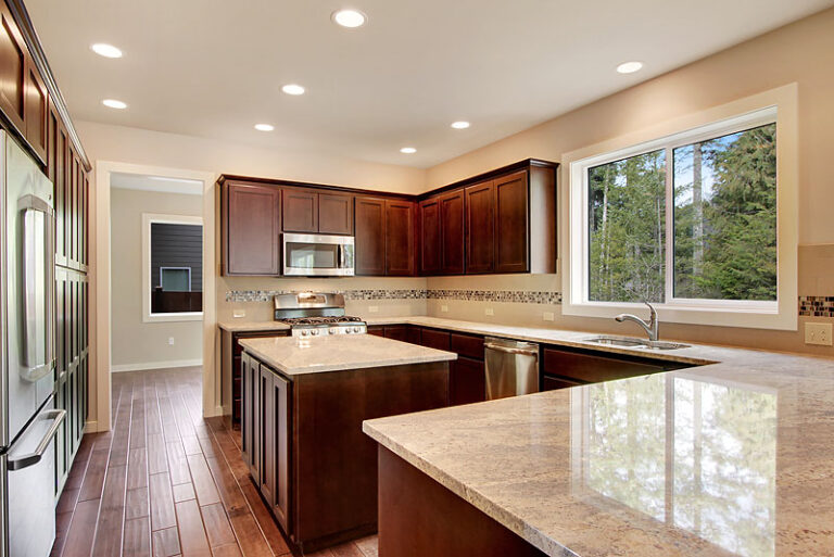 A spacious kitchen with dark wooden cabinets, stainless steel appliances, granite countertops, a large window with a forest view, and hardwood flooring.