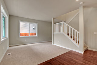 This image shows an empty room with beige walls, carpeted and hardwood floors, a large window, and a white staircase railing to the right.