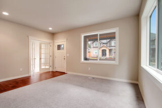 Empty room with beige walls, large window with a view of houses outside, hardwood floor entryway transitioning to gray carpet, and white doors.