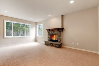 The image shows an empty room with beige carpeting, a stone fireplace with a lit fire, large windows displaying a forest view, and white walls.