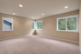 This image shows an empty room with beige walls, carpeted flooring, recessed lighting, and three windows looking out to greenery.