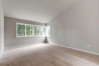 Empty room with beige carpet, white walls, and a set of three windows allowing natural light, overlooking green trees outside, suggesting a peaceful interior space.