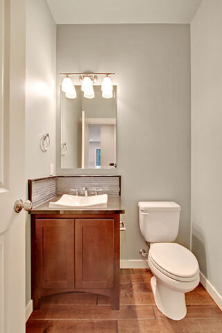 A small, modern bathroom with a toilet, wooden vanity cabinet, vessel-style sink, and a five-light fixture above a mirror on a light green wall.