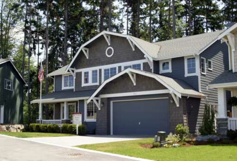 A two-story residential house with gray siding, white trim, a double garage door, and an American flag displayed. Lush green trees and a clear sky are visible.