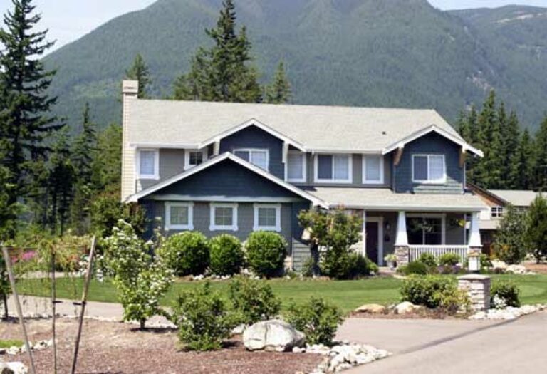  A two-story suburban house with a green exterior and white trim. Landscaped garden, mountains in the background, clear sky, and a paved driveway. 