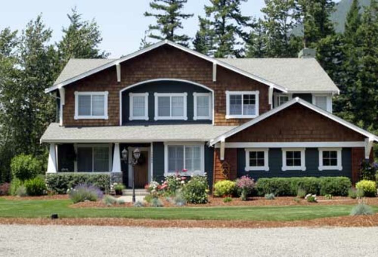 A two-story suburban house with dark trim and a green roof. Landscaped with flowers, shrubs, and a well-manicured lawn. Gravel driveway in front. Clear sky.