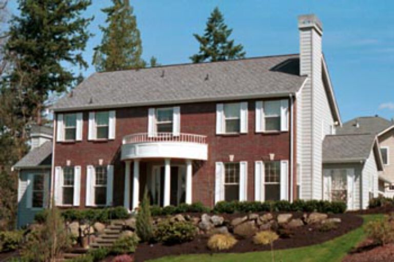 Two-story suburban house with a red brick and white siding exterior, large front windows, a curved balcony, a prominent chimney, and green landscaping.