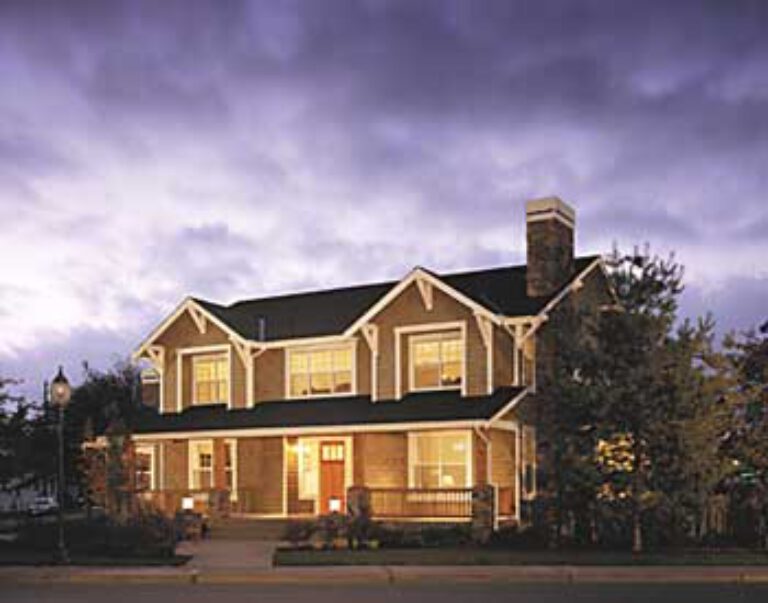  A two-story house with a gabled roof, prominent chimney, and porch illuminated by warm light. Dusk setting with vibrant purple clouds overhead. 