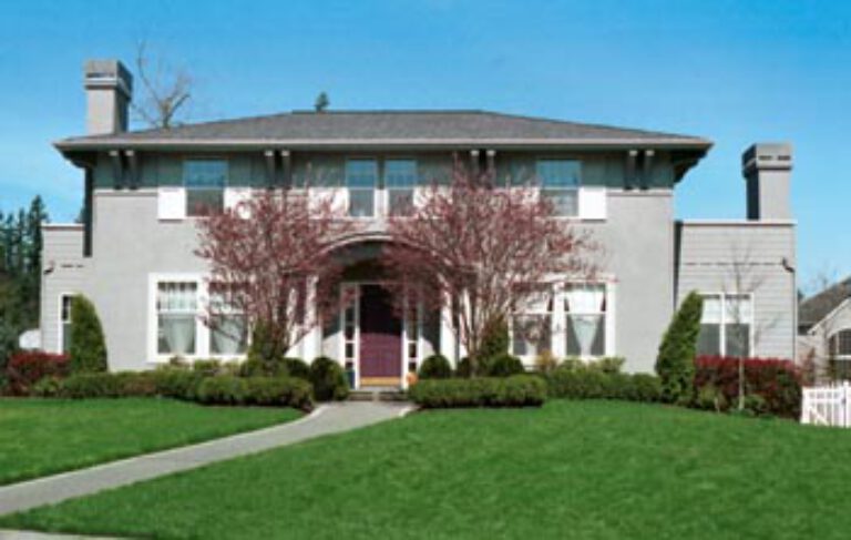 This is a symmetrical two-story house with a gray roof, beige walls, and a central arched entrance. It's surrounded by a manicured lawn under a clear sky.