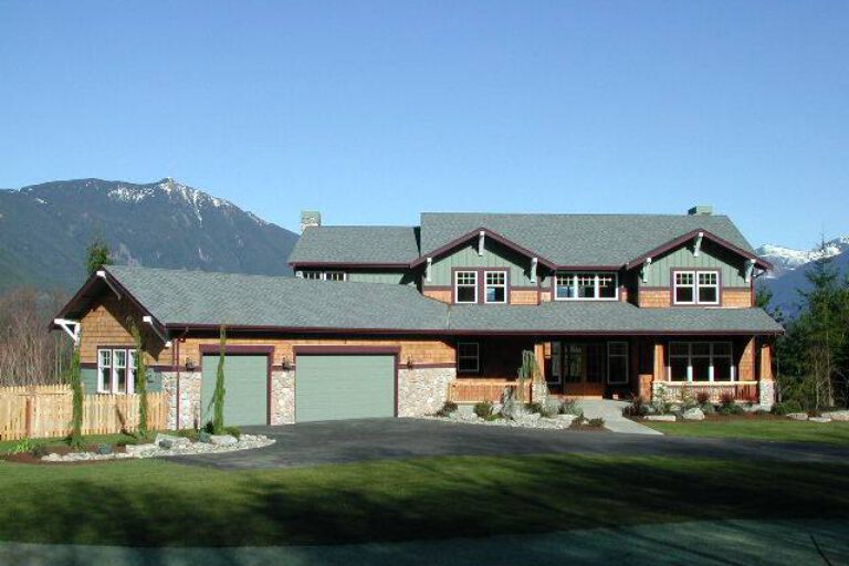 A spacious two-story house with a green garage door, wooden accents, multiple windows, set against a backdrop of mountains and a clear blue sky.