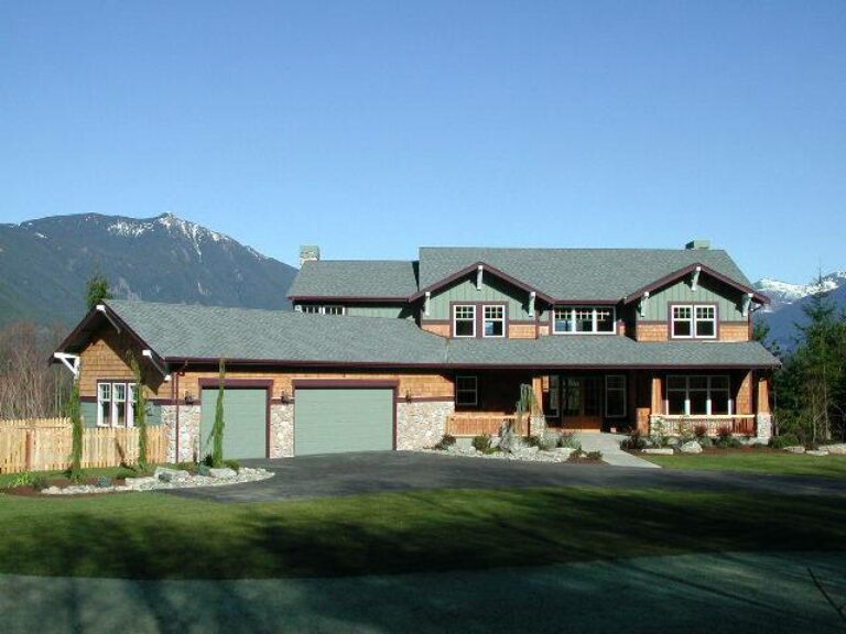  Large two-story house with green roof and garage doors, surrounded by a wooden fence, set against a backdrop of mountains under a clear blue sky. 