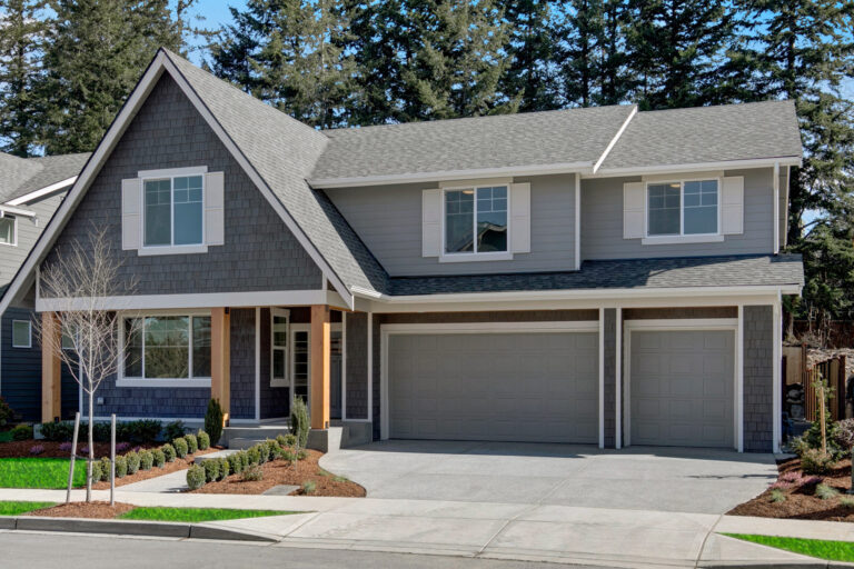 This image features a two-story suburban house with a gray exterior, white trim, dual garage doors, and a landscaped yard with evergreen trees.