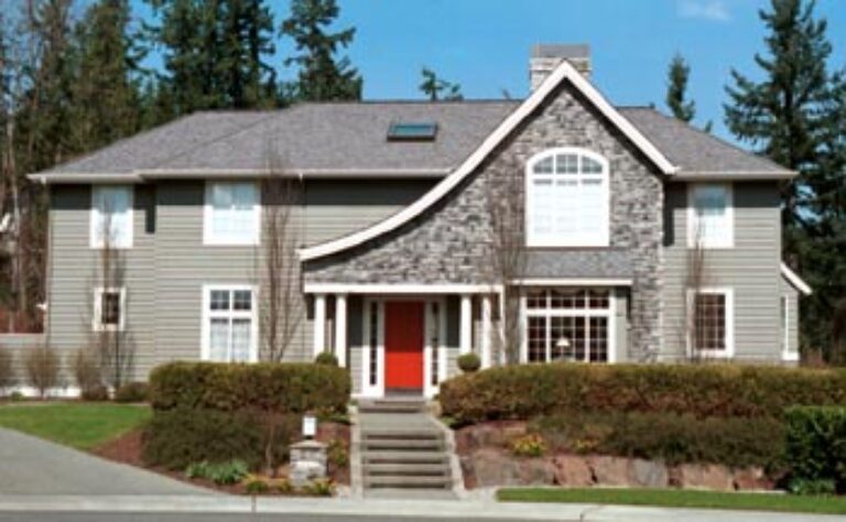 This image shows a two-story suburban house with gray siding, white trim, a red door, and a gable roof surrounded by a manicured lawn and clear skies.