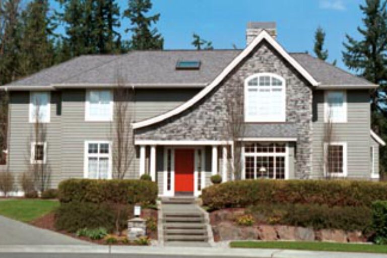 This image shows a two-story suburban house with gray siding, white trim, a red door, and a gable roof surrounded by a manicured lawn and clear skies.