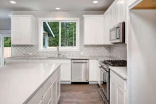 A modern kitchen with white cabinetry, herringbone backsplash, stainless steel appliances, and a view of green trees through the window.