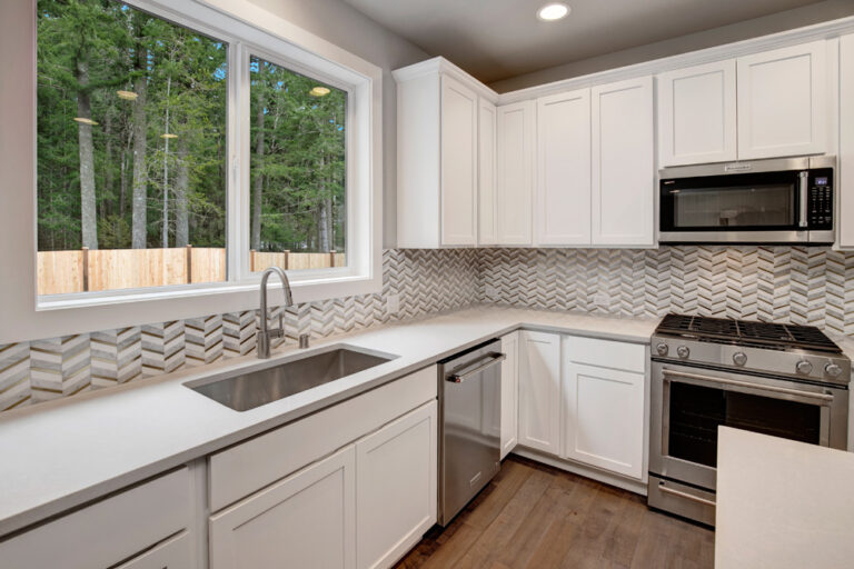 The image shows a modern kitchen with white cabinets, stainless steel appliances, a herringbone backsplash, and a forest view from the window.