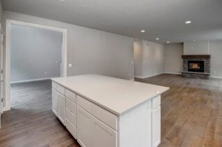 Modern empty room with hardwood floors, a white kitchen island, gray walls, recessed lighting, and a stone fireplace with a lit fire.