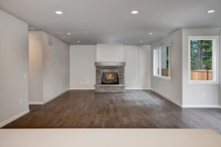 A spacious, empty room with hardwood floors, white walls, recessed lighting, a stone fireplace, and windows looking out to trees.