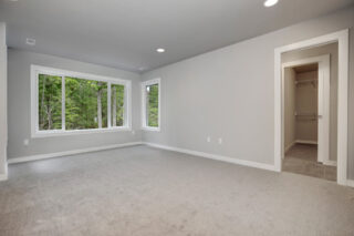 The image shows an empty room with gray walls and carpet, two large windows with a forest view, recessed lighting, and an open closet.