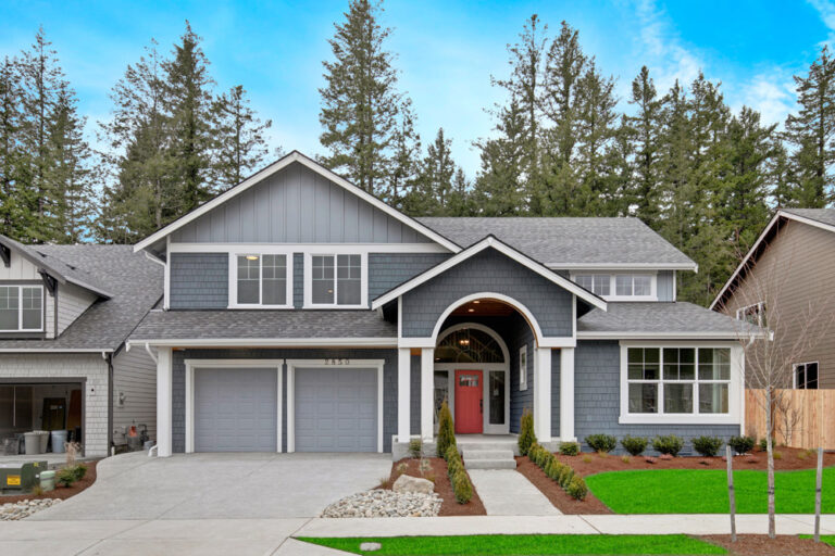  A two-story suburban house with gray siding, white trim, a covered entry archway, red door, and a double garage, set against a backdrop of tall trees. 