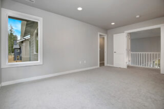 An empty room with gray carpeting, white walls, recessed lighting, a large window, a white door, and an open passageway to a railed area.