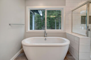 Modern bathroom interior featuring a standalone white bathtub, a window with a forest view, gray tiled floor, and white subway tiled walk-in shower area.